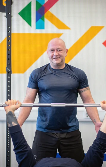 Student performing a bench press while another student spots them in a colourful training area. Student performing a bench press while another student spots them in a colourful training area.
